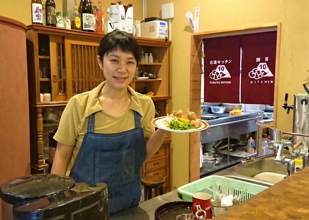 飲食店の厨房内で、デニムのエプロンを付けた女性店員が揚げ物と生野菜が盛られた皿を手に微笑んでおり、背景には「古湯キッチン10」と記された暖簾や酒瓶が並ぶ食器棚が見える、アットホームな雰囲気の調理場の写真