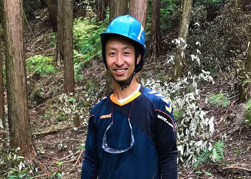 杉木が立ち並ぶ深い森の中で、青いヘルメットを被り首から保護メガネを下げた笑顔の男性が、紺色のスポーツウェアを着用してカメラを見つめて立っている、林業や屋外作業に従事している様子を捉えた写真