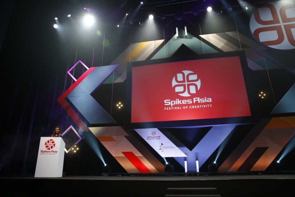 スポットライトが照らす暗いイベントステージで、中央の巨大スクリーンに「Spikes Asia FESTIVAL OF CREATIVITY」の赤いロゴが表示され、左側の演台にはロゴと同じマークが描かれた女性が立つ華やかな授賞式会場の写真