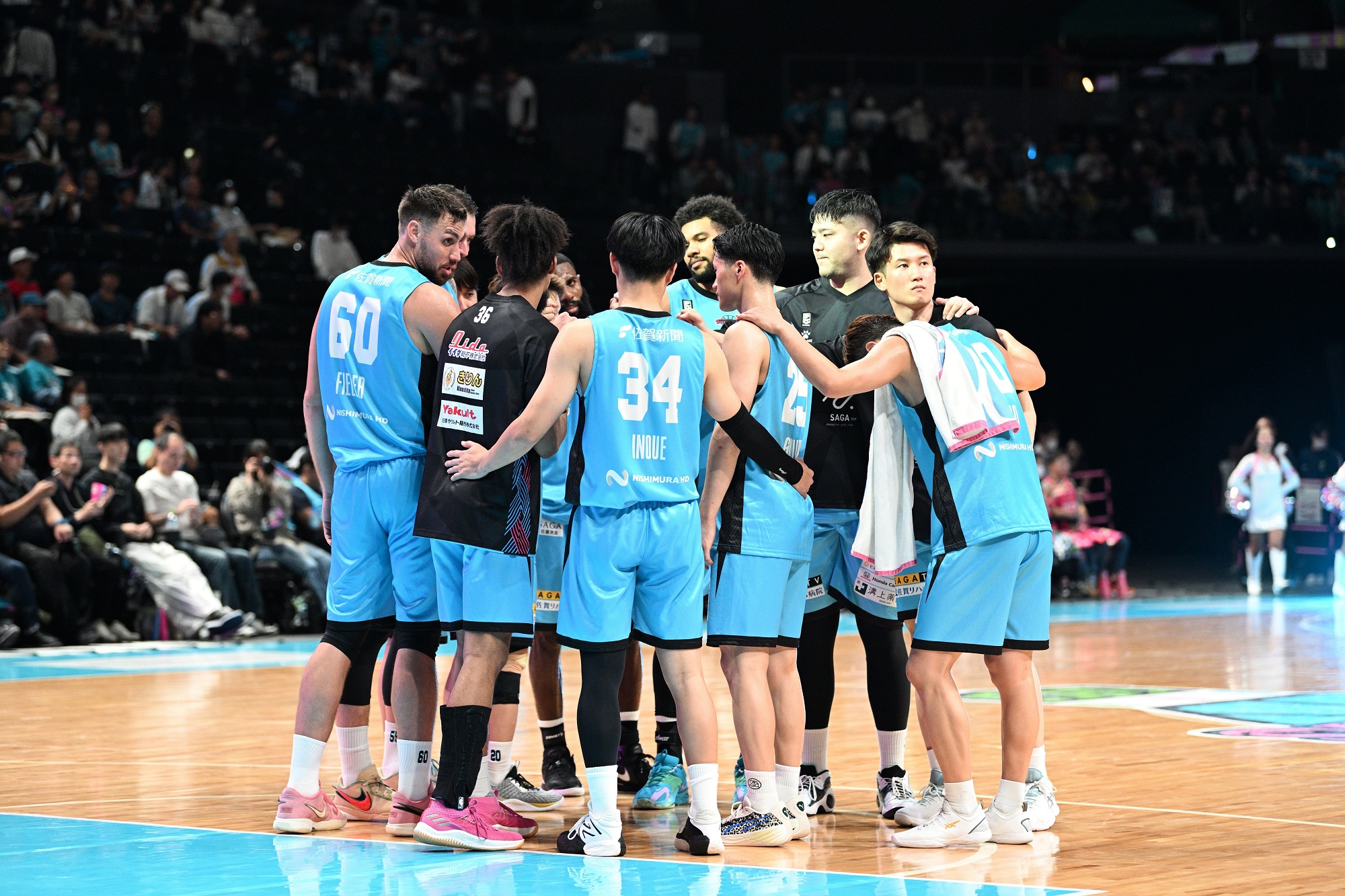 水色のユニフォームを着用したバスケットボールチームの選手たちが、試合会場のコート中央で円陣を組み、互いの肩に手を置きながら真剣な表情で戦略を練ったり鼓舞し合ったりしている写真