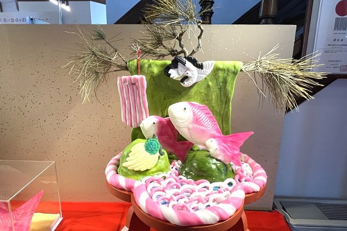 松の枝の下に、砂糖などで作られた3匹の紅白の鯛の精巧な細工が緑色の台の上に華やかに盛り付けられた、伝統的な祝い菓子「寿賀台」が展示されている様子を捉えた写真