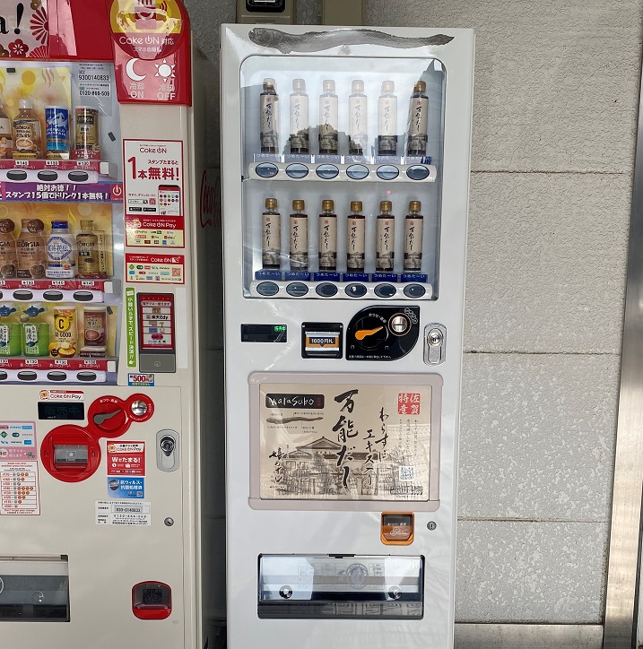 上部には細長い魚のイラストが描かれ、中央の大きなパネルには商品の特徴を説明する筆文字のテキストや建物の外観が描かれている、「わらすぼエキス入りの万能だし」が販売されている白い自動販売機を正面から捉えた写真