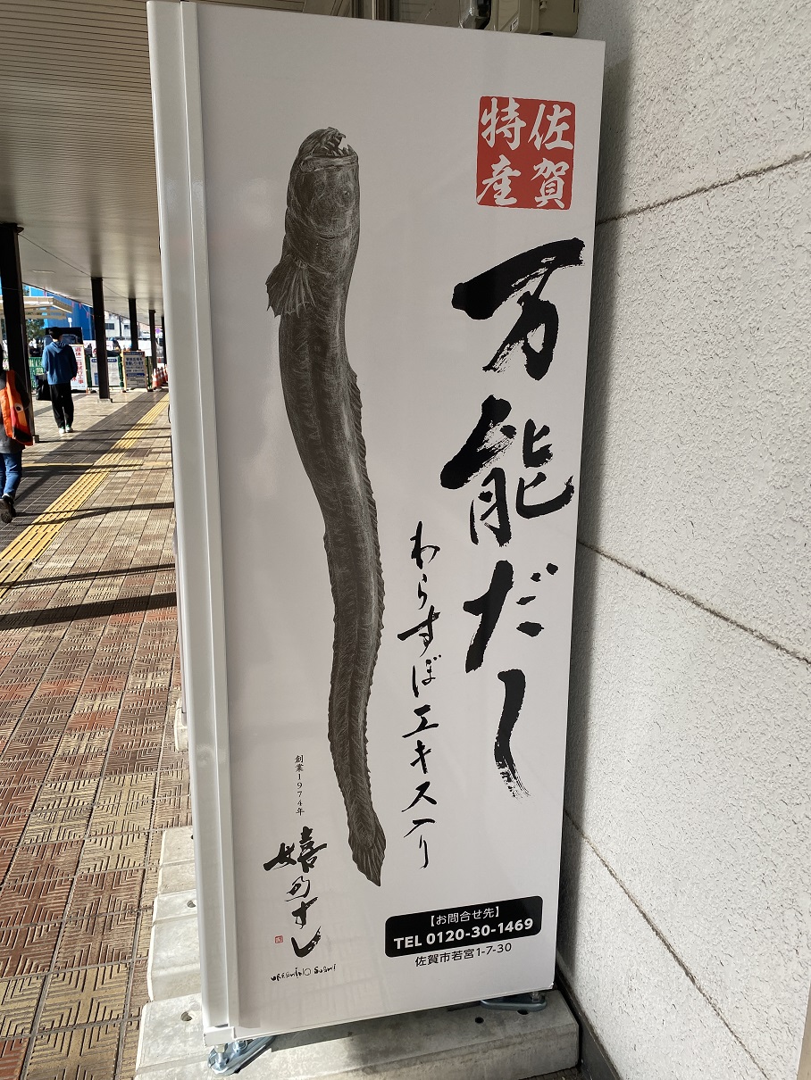 大きく口を開けたワラスボのイラストと「万能だし わらすぼエキス入り」という筆文字のテキストが書かれ、右上には「佐賀特産」の赤い印影が配置された、白い自動販売機の側面を捉えた写真