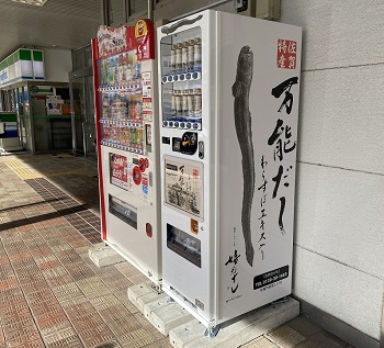 屋外の店舗前に設置された、佐賀特産「わらすぼエキス入り 万能だし」の大きなイラストと商品名が側面に描かれた白い自販機と、その隣に並ぶ一般的な飲料自販機がタイル敷きの歩道に佇む様子を捉えた写真