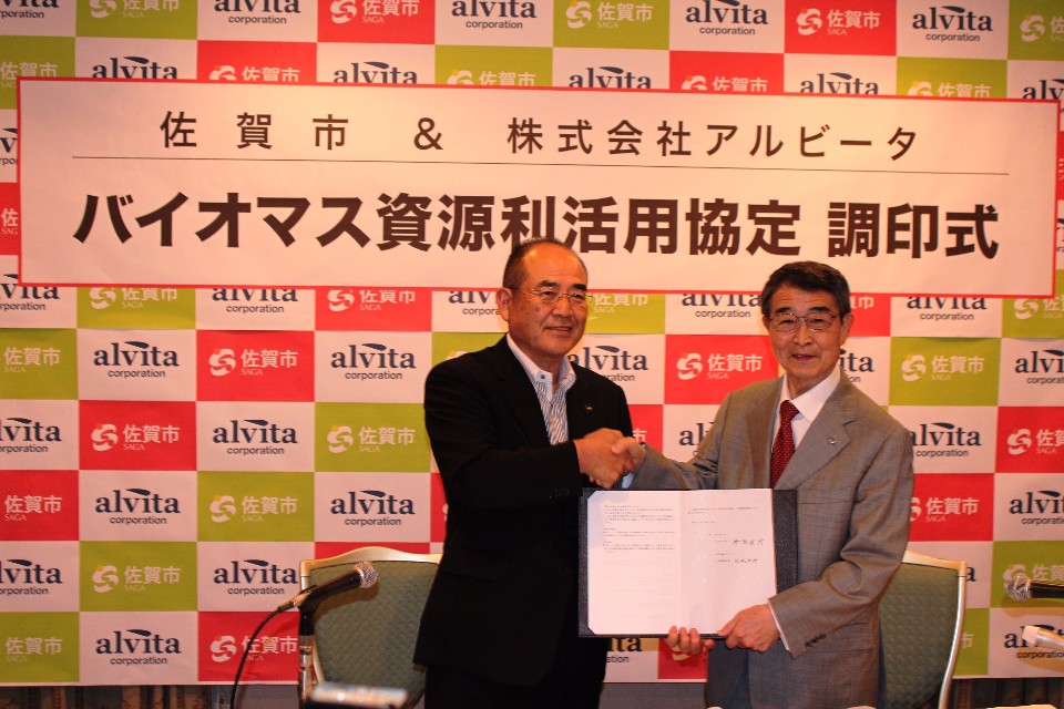 バイオマス資源利活用協定の調印式でスーツ姿の市長と代表の男性二人が協定書を示しながら握手を交わし、社名入りの背景幕と壇上のテーブルや椅子が並ぶ写真