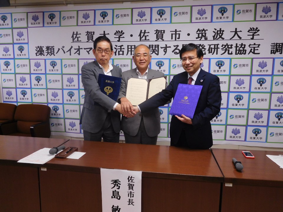 佐賀大学・佐賀市・筑波大学の協定調印式で男性三人が協定書や資料を掲げつつ握手を交わし、背景に各機関のロゴが並ぶ写真