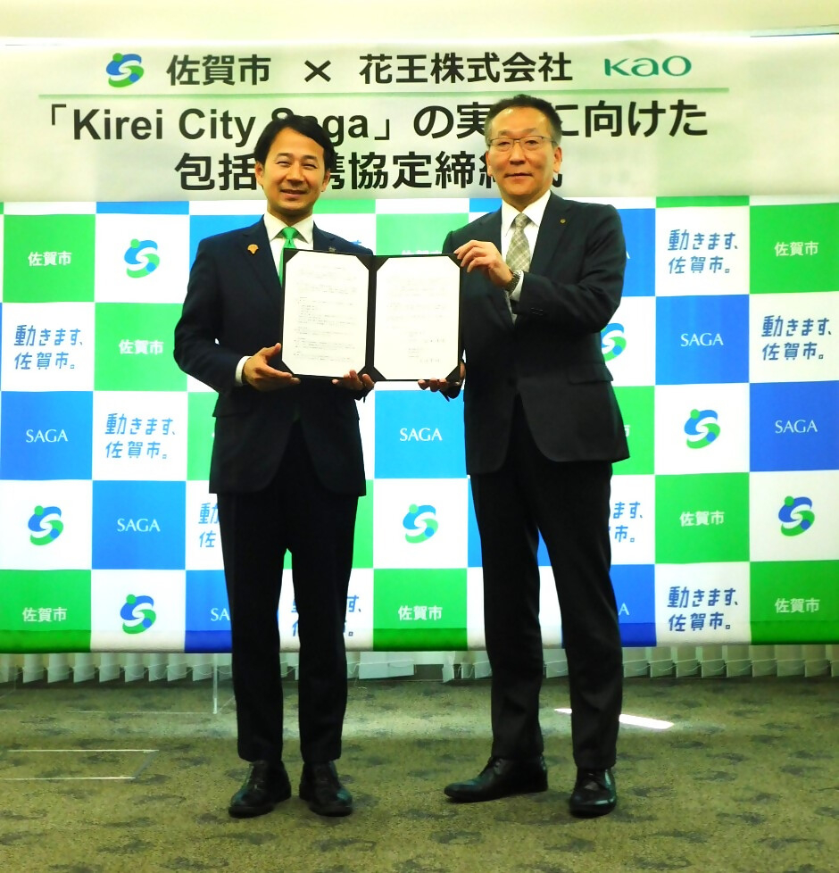 佐賀市と企業の協定締結式で二人が前方で協定書を並べて掲げ背景に記者会見用パネルが並ぶ写真