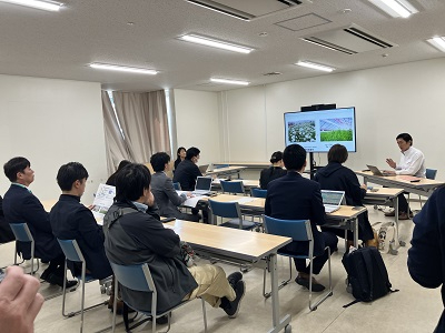 教室内で参加者が机に着席し前方の大型モニターを囲んで発表者の説明を熱心に聞く様子の写真