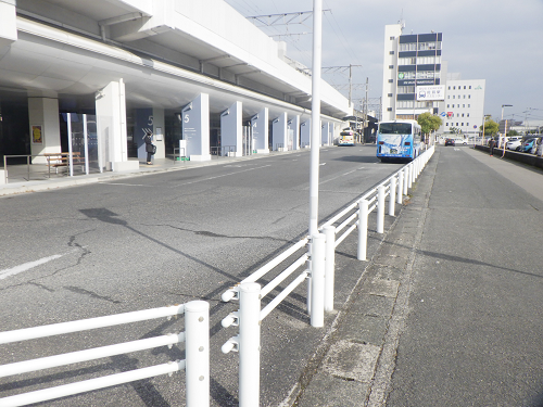 高架下の広い車道沿いに白い柵が続き、奥には青い路線バスが停車する静かな街並みを写した写真
