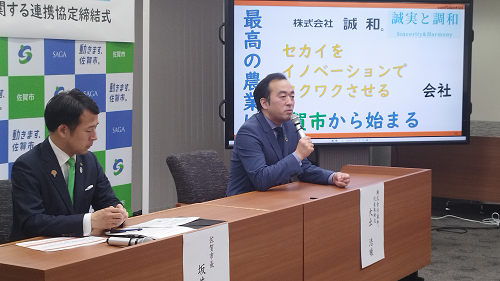 「セカイをイノベーションでワクワクさせる」と記されたモニターを背に、マイクを握って話す株式会社誠和 大出 浩睦代表取締役と、机に座り資料に目を落とす佐賀市長の写真
