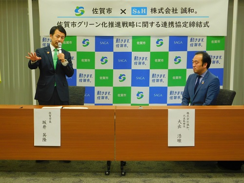「佐賀市グリーン化推進戦略に関する連携協定締結式」の看板の前で、マイクを持ち身振り手振りを交えて話す坂井英隆佐賀市長と、その隣で真剣な表情を浮かべて着席している株式会社誠和の大出浩睦代表取締役の写真