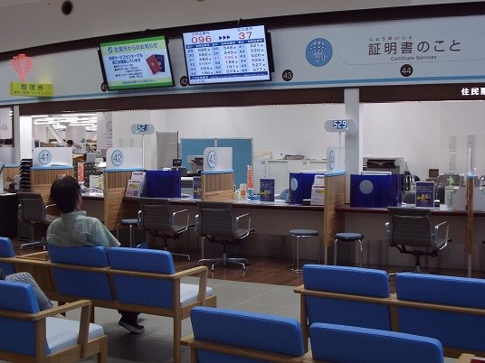 市役所内に並んだ青い待合席の正面に、複数の窓口があり、その上部に番号案内表示モニターが設置されている写真