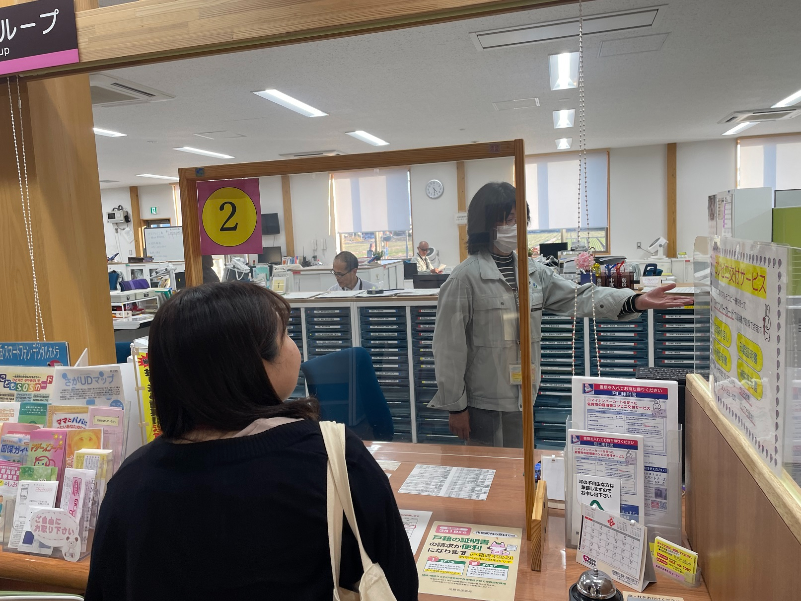 案内用のパンフレットや掲示物が並ぶ飛沫防止パネルが設置された役所の窓口で、作業服姿の職員がカウンター越しに来庁者の女性へ向けて手を差し出しながら説明を行っている写真