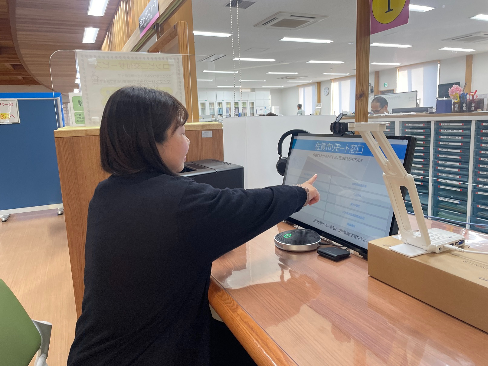 黒い服を着た来庁者の女性が「佐賀市リモート窓口」と表示されたタッチパネル式のモニターを操作しており、デスクにはスピーカーフォンやカメラが設置されている写真