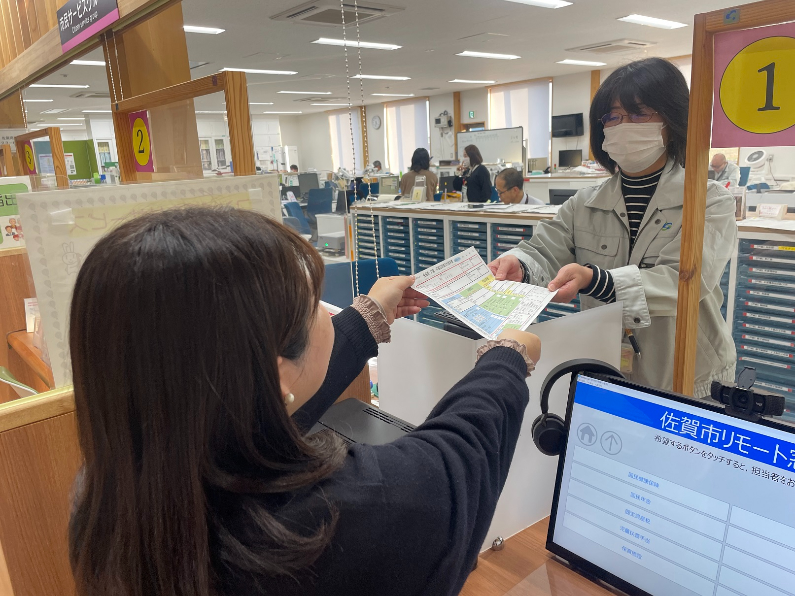 来庁者の女性が「佐賀市リモート窓口」と表示されたモニターの横から、職員へ記入した申請書を手渡している様子の写真
