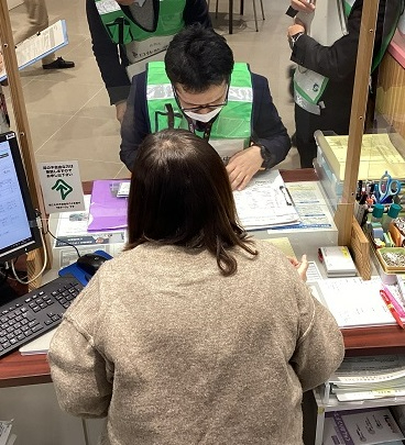 窓口カウンター越しに来訪者が書類を確認しながら職員と手続きを進め周囲にパソコンや筆記具が並ぶ様子を示した窓口体験調査の様子写真