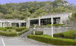 緑豊かな山を背景に、石造りの外壁と大きなガラス窓を備えた低層の建物が階段と植え込みに囲まれている、つくし斎場の外観写真