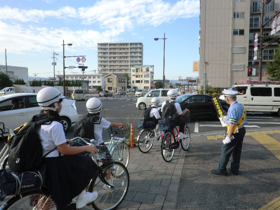 交差点近くの歩道でヘルメットを着用した複数の学生が自転車に乗って信号待ちをしており、横には黄色い腕章を付けた交通安全指導員が立っている写真