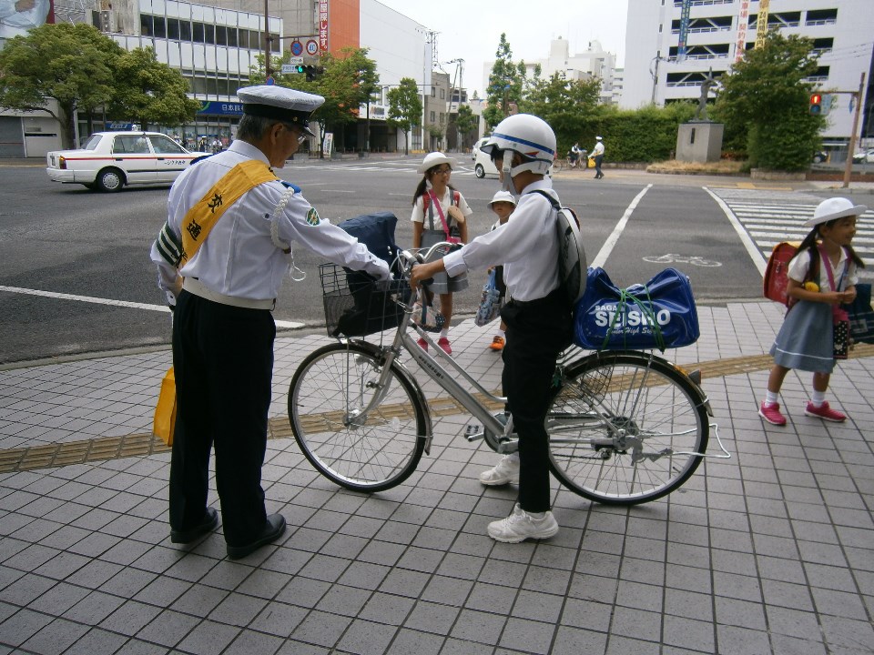 横断歩道と車道が広がる市街地の歩道上で制服姿の交通安全指導員が自転車に乗るヘルメット着用の児童に声をかけて街頭指導を行っている様子の写真