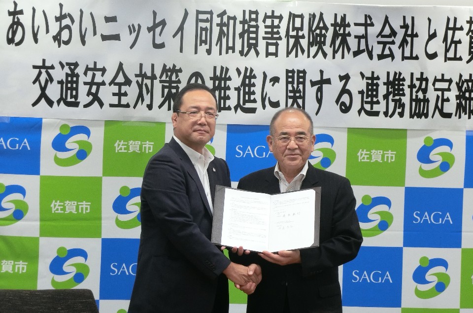 背景に「交通安全対策の推進に関する連携協定締結」と大きく書かれた横断幕が掲げられ、佐賀市のロゴ入りパネルの前で二人の男性が協定書を広げて握手している写真