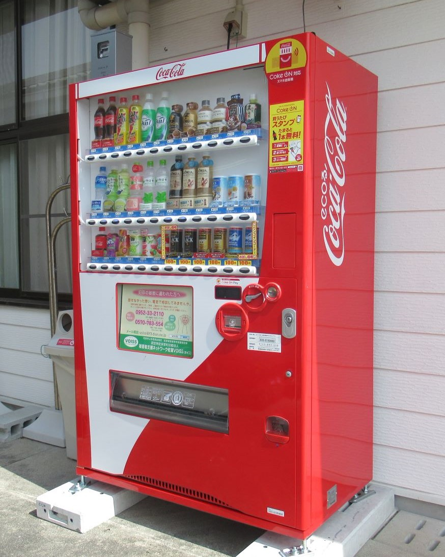 赤と白を基調としたコカ・コーラの自動販売機で、上段にはペットボトル飲料、下段には缶コーヒーやジュースが並び、側面にブランドロゴが大きく表示された屋外設置の写真