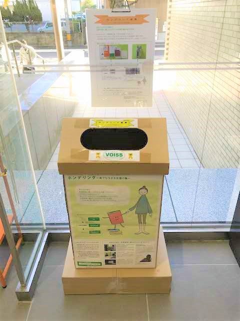 ガラス張りの室内に設置された段ボール製の回収ボックスで、正面には赤い本を持つ人物のイラストと案内文、上部には「VOISS」と書かれたラベルが貼られた写真