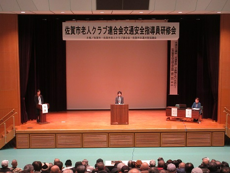 大きな講堂で開催された交通安全指導員研修会の壇上に登壇者が立ち、客席の参加者が真剣に聴講する様子を写した写真