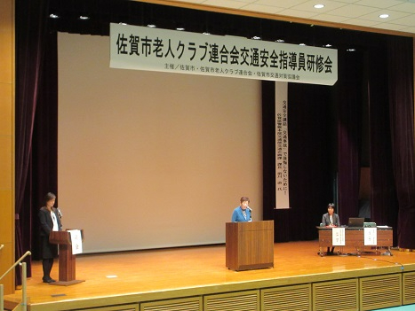 大きな講堂の壇上で交通安全指導員研修会の演台に登壇者が立ち両脇の係員とともに進行する様子を捉えた写真