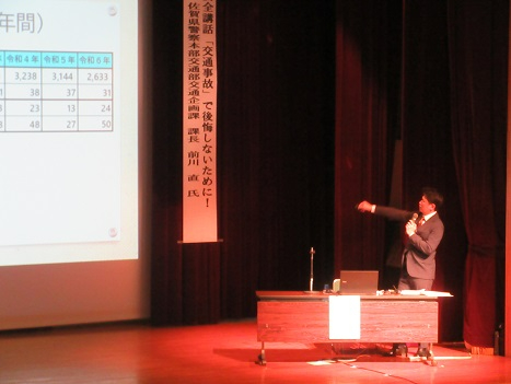 講堂の壇上で講師がマイクを手にスクリーンの統計表を指し示しつつ交通安全の要点を丁寧に解説する様子の写真