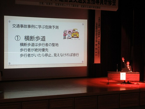 暗い講演会場のステージ上で、「交通事故事例に学ぶ危険予測 1横断歩道」というスライドがスクリーンに投影されており、その横のデスクで挙手しながら解説を行う講師と、歩行者優先を訴えるイラストが表示されている講習会の様子を捉えた写真
