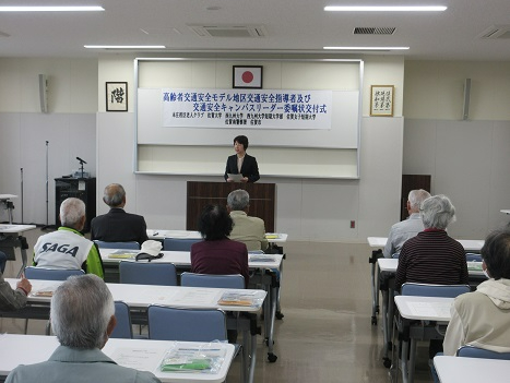 会議室で壇上の講師が参加者たちに向けて話をしている様子を室内の後方から撮影した写真