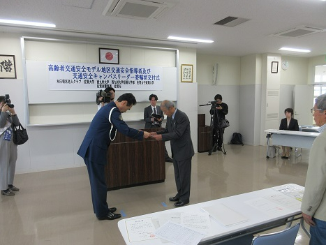 委嘱状交付式の会場で制服姿の男性が高齢の男性に委嘱状を丁寧に手渡す様子を周囲の関係者が見守っている写真