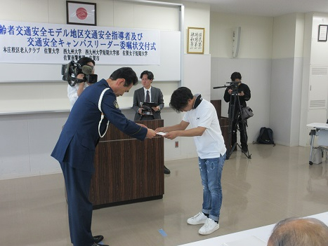 委嘱状交付式で紺の制服姿の男性が白いシャツの若い男性に委嘱状を丁寧に手渡し周囲の関係者が静かに見守る様子の写真