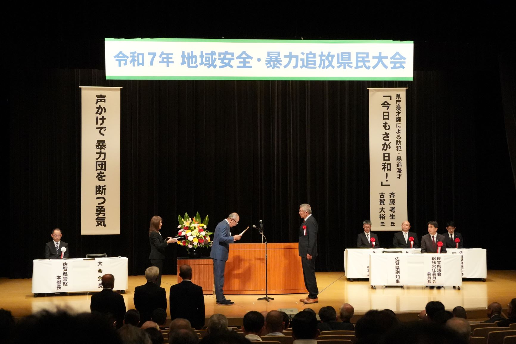 「令和7年 地域安全・暴力追放県民大会」の看板が掲げられた舞台上で、スーツ姿の受賞者と関係者の男性が向かい合って立ち、授賞式が行われている様子を写した写真