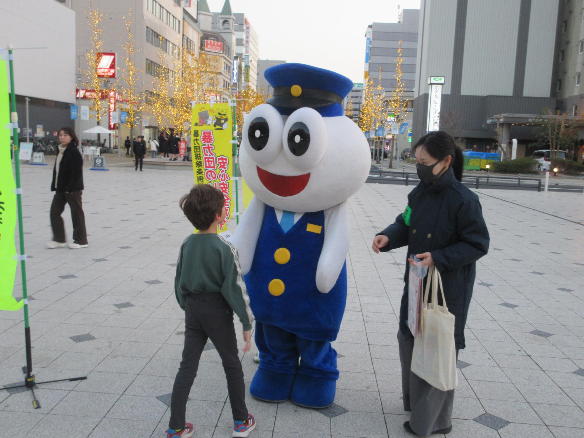 大きな丸い瞳が特徴的な佐賀県警キャラクターごろうくんと啓発運動に参加している女性が、男の子と交流している街頭での街頭キャンペーンの様子を写した写真