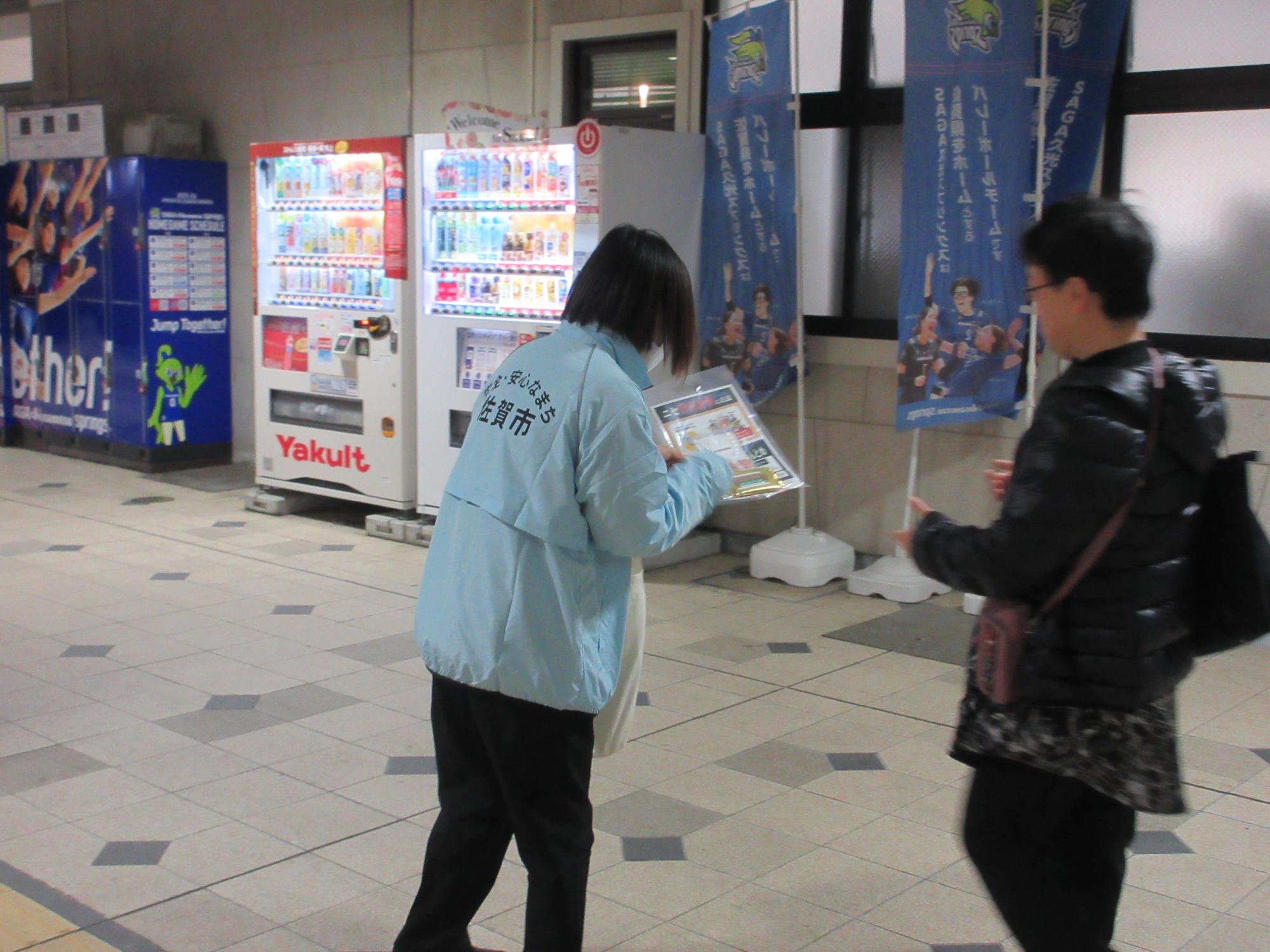 「佐賀市」の文字が入った水色のジャンパーを着た啓発活動参加者の女性が、歩行者に防犯啓発のチラシが入った袋を手渡そうとしているキャンペーン活動の様子を写した写真