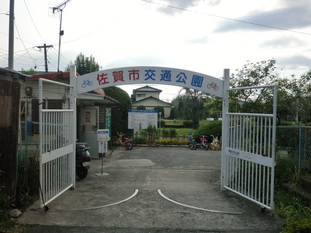 白い門に「佐賀市交通公園」と青赤文字で書かれ、自転車のイラストが両端に描かれた入口の奥に案内板と複数の子供用自転車が並ぶ緑豊かな公園の写真