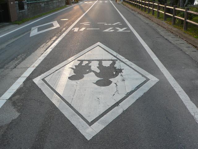舗装された道路の中央に児童のシルエットが描かれた大きな白い菱形マークがあり、周囲には白線で区切られたスクールゾーンの表示とガードレールが続く通学路の写真