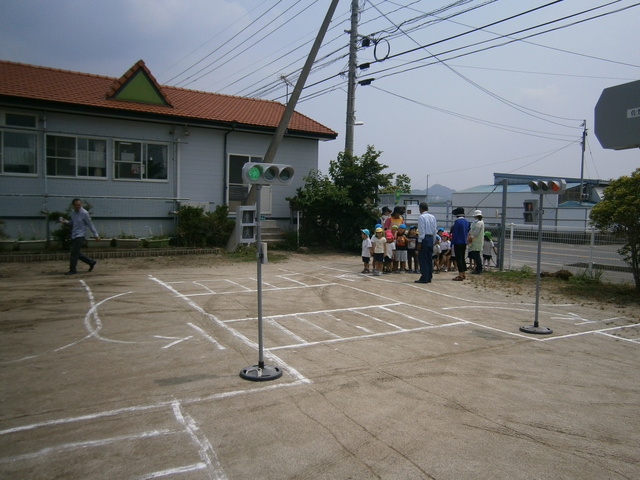園庭の一角に白線で交差点や横断歩道が描かれ、簡易信号機が設置された場所で、複数の子どもと大人が集まり交通安全指導を受けている様子の写真