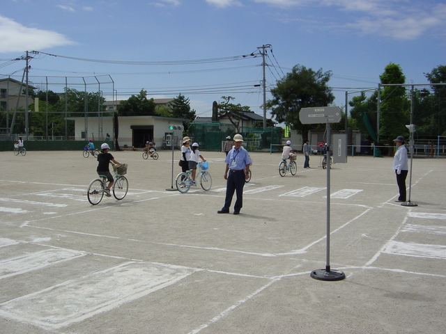 白線で交差点や横断歩道が描かれた広い校庭で、複数の子どもが自転車に乗り、数名の大人が指導する交通安全教室の様子の写真