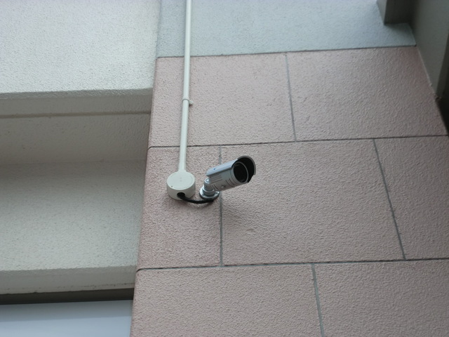 壁面に取り付けられた銀色の防犯カメラが、ピンク系タイルの外壁と白い配線ボックスに接続されている写真
