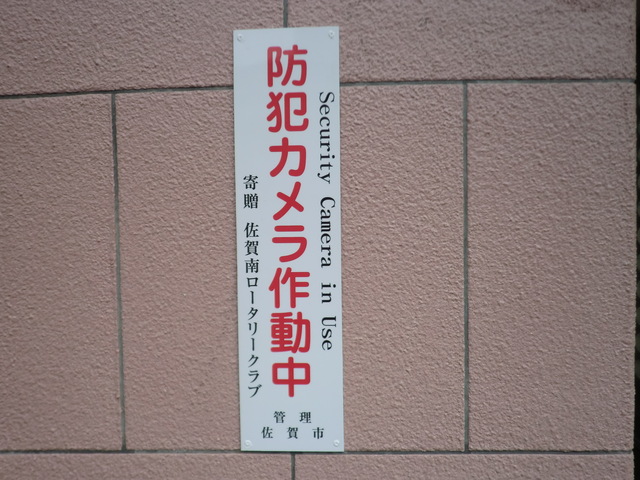ピンク色のタイル壁に、赤文字で「防犯カメラ作動中」、黒文字で寄贈者名や管理者名、英語で「Security Camera in Use」と記載された縦長の白い看板が取り付けられている写真