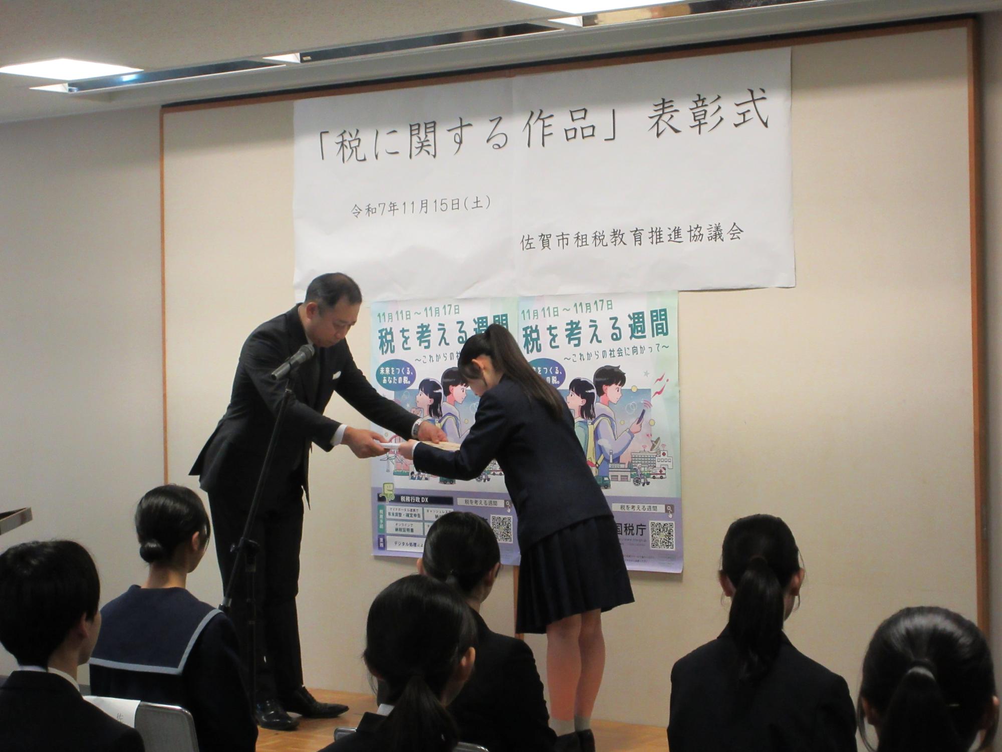 「税に関する作品」表彰式と書かれた幕の前で、左に男性、右に女子生徒が向かい合って立ち賞状を受け取っている表彰式の様子の写真