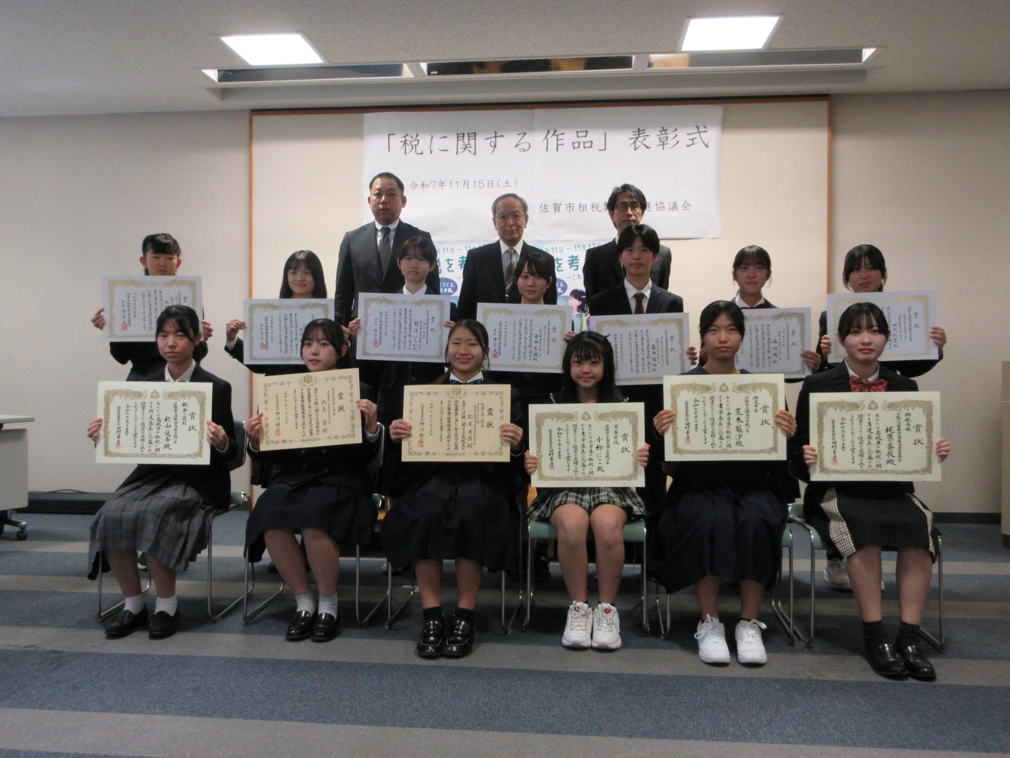 「税に関する作品」表彰式と書かれた幕の前で、賞状を掲げた制服姿の生徒たち13名が2列に並び、後列にスーツ姿の男性3名が並んだ集合写真