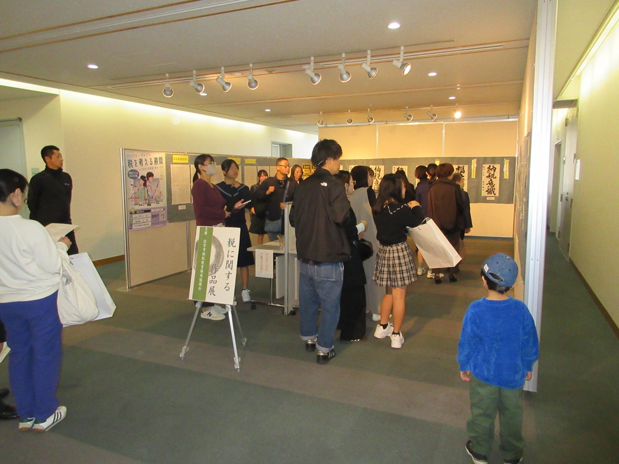 「税に関する作品展」と書かれた展示会場で、壁に並んだ習字作品・作文などを鑑賞する多くの来場者たちの写真