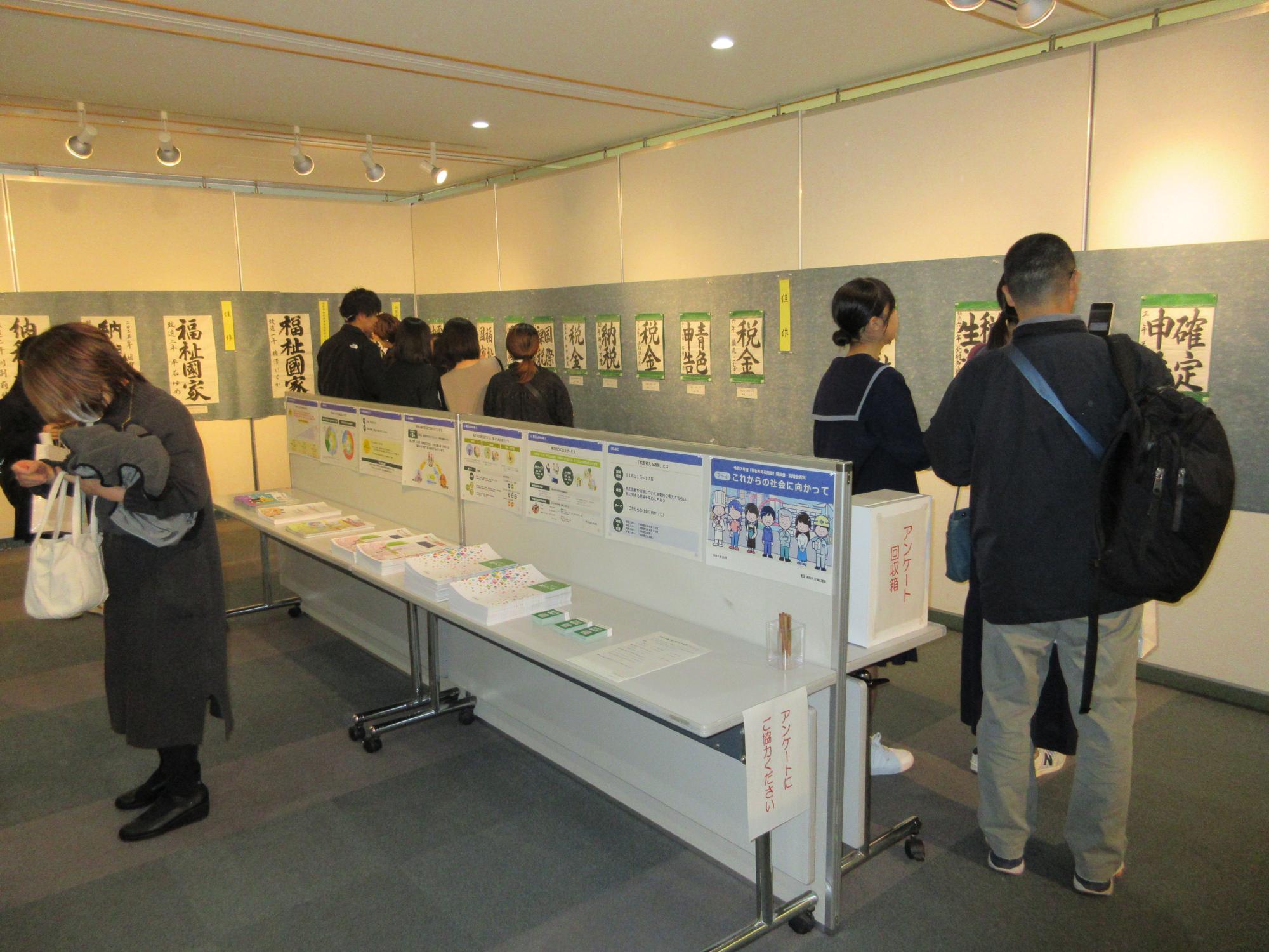 「展示会場で、壁に並んだ習字作品を鑑賞する来場者たちや、手前のテーブルに置かれたパンフレット、アンケート回収箱などが並ぶ屋内の様子を捉えた写真