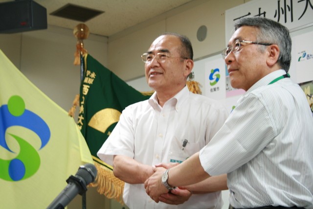 「西九州大学との協定締結式」において、秀島敏行市長と福元裕二学長が、佐賀市の旗や大学の旗を背景に握手を交わしている様子を写した写真