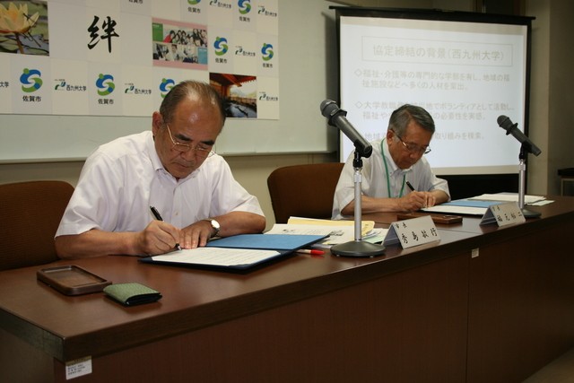 「西九州大学との協定締結式」において、秀島敏行市長と福元裕二学長が並んで座り、手元の青いファイルに署名を行っている写真