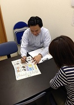 机を挟んで向かい合った男性担当者と女性が、広げた資料を指し示しながら静かな雰囲気で説明を進めている様子の写真