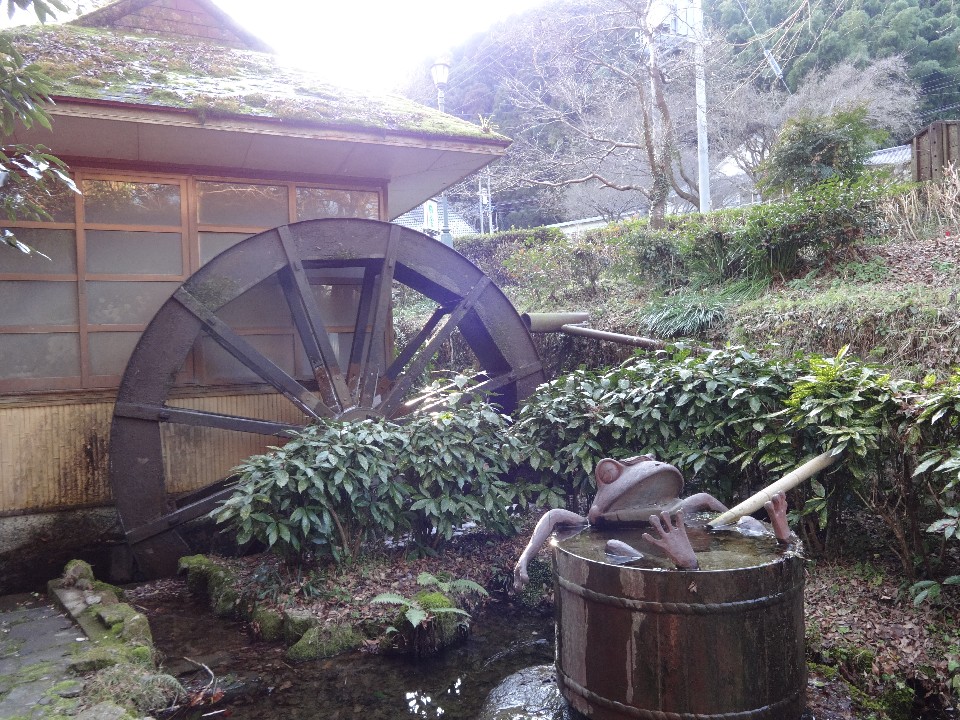 苔むした水車小屋のそばで小川に置かれた樽からユーモラスな蛙の像が顔を出す情緒ある風景の写真